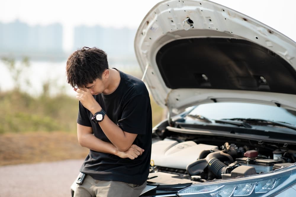 Concerned young Asian man inspecting his car after a breakdown. Broken-down vehicle on the road, requiring emergency service assistance.