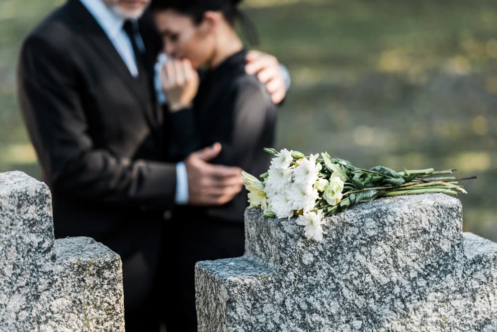 Close-up of a bouquet resting on a tombstone with a man tenderly embracing a woman in the background.