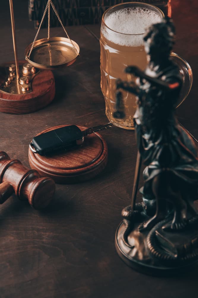 Statue of justice, scales, gavel, car key, and a mug of beer on a table, representing legal consequences of drunk driving.