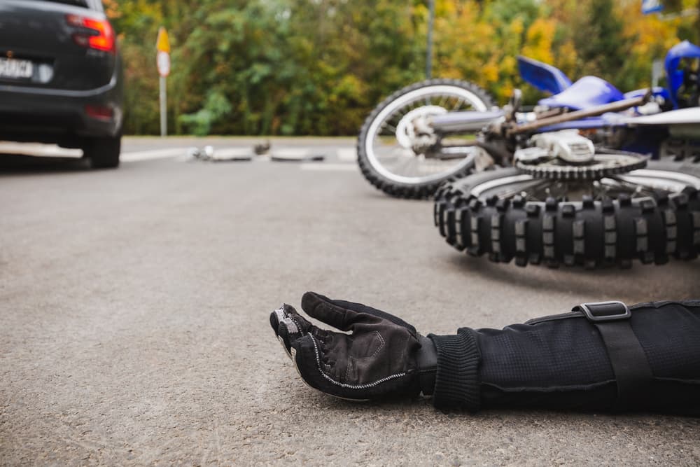A motorcycle accident scene with a fallen bike and a rider's gloved hand on the pavement, car in the background.