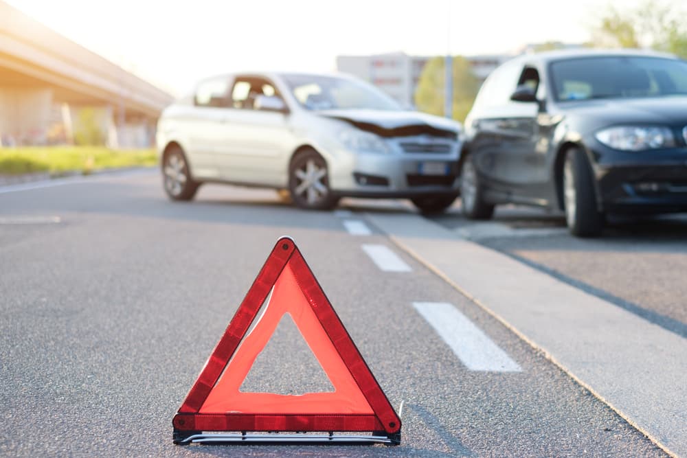 A red warning triangle on a road with two cars crashed in the background, indicating a recent traffic accident.