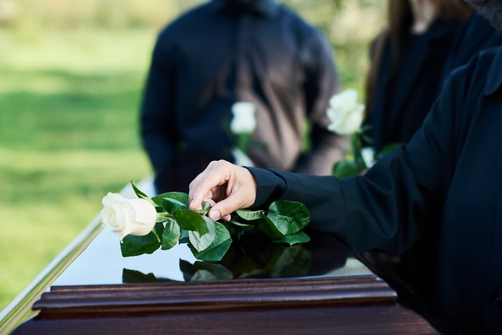 Mature woman in mourning attire delicately placing a white rose atop a closed coffin lid, captured in a poignant moment as she stands before the camera amidst a backdrop of other mourners.
