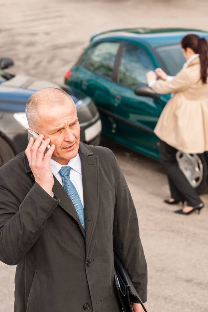 A man in a suit talking on a phone looks concerned, with a woman examining a car in the background.
