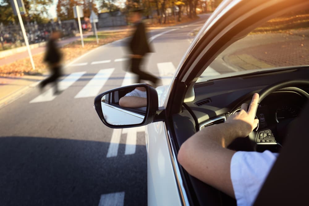 A driver at a crosswalk watches two blurred pedestrians crossing the street from the car's side mirror.