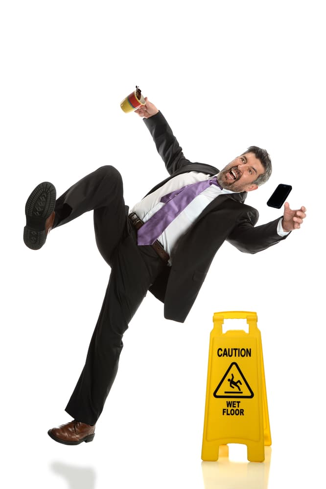 Latino businessman slipping beside wet floor caution sign on white backdrop, emphasizing workplace safety and potential hazards.