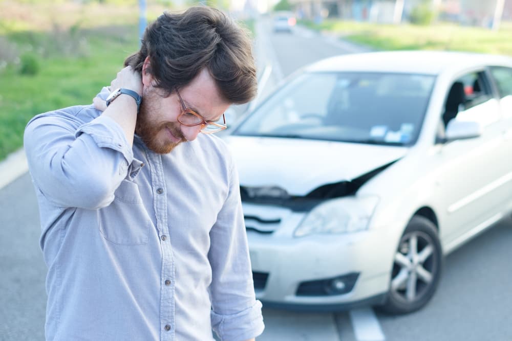 Man feeling neck pain after a minor car accident, standing on the roadside with damaged vehicles in background.