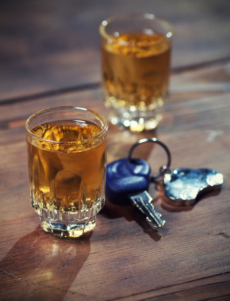 Two glasses of whiskey on a wooden surface next to a set of car keys, suggesting the dangers of drinking and driving.