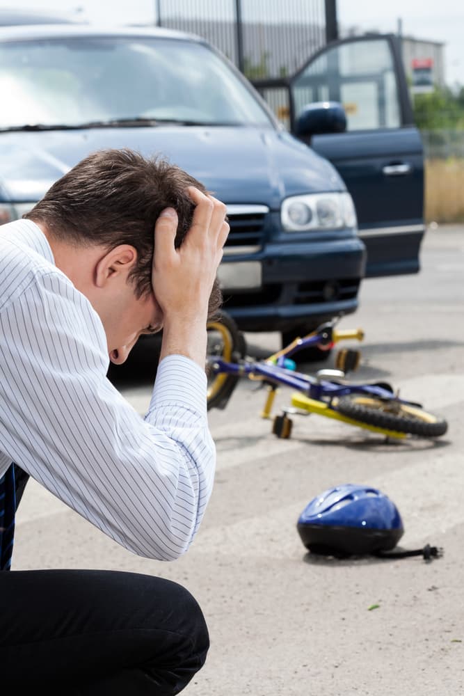 Distressed man crouching on pavement with hands on head near a fallen bicycle and helmet, with a car in the background.