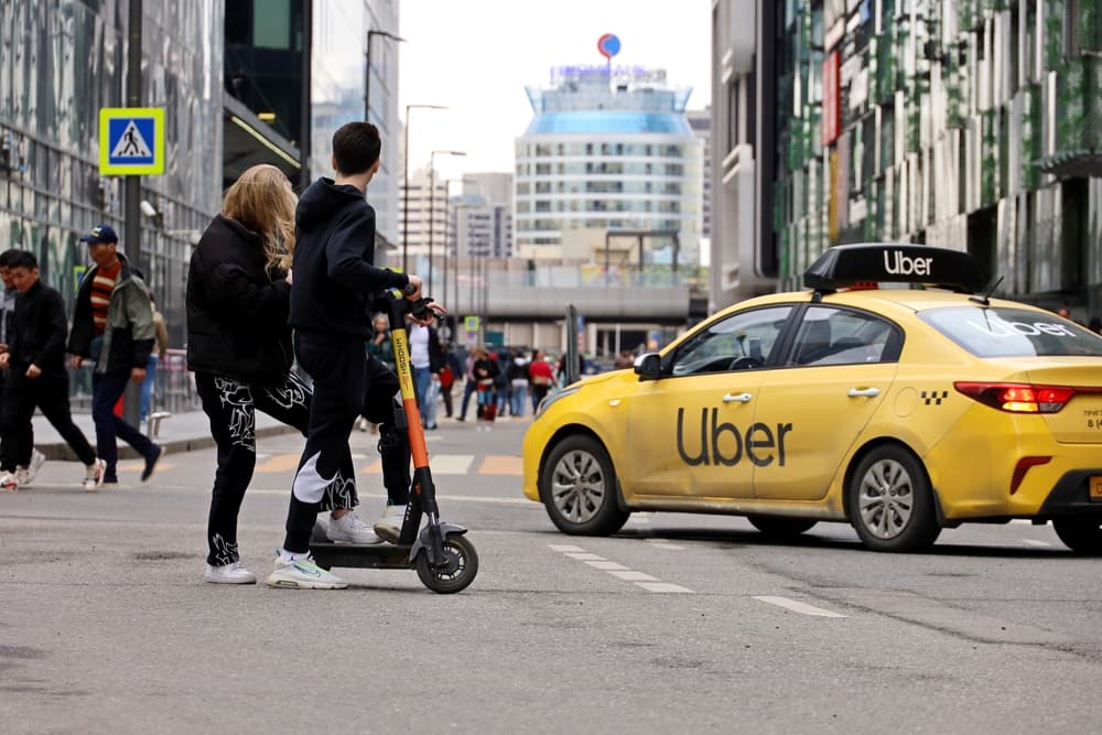 Two individuals riding an electric scooter crossing the street beside a stationary yellow Uber car in a busy urban setting.