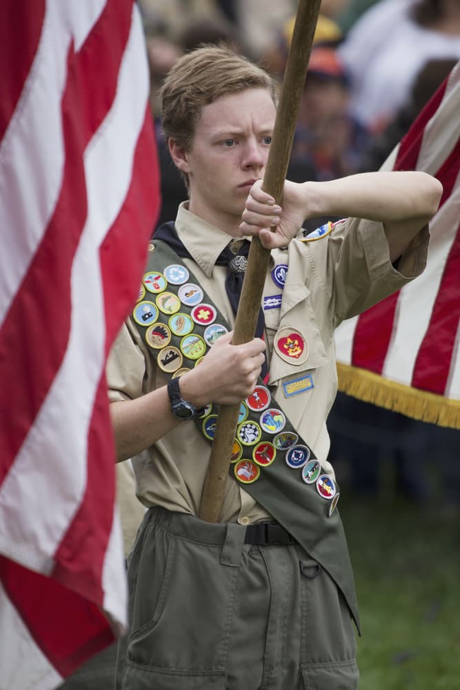 A Boy Scout in uniform solemnly holding a flagpole with numerous merit badges displayed.