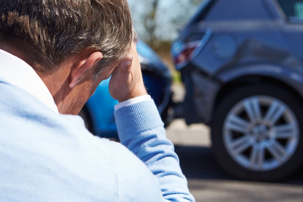 A man appears stressed, holding his head, with a damaged car visible in the background.