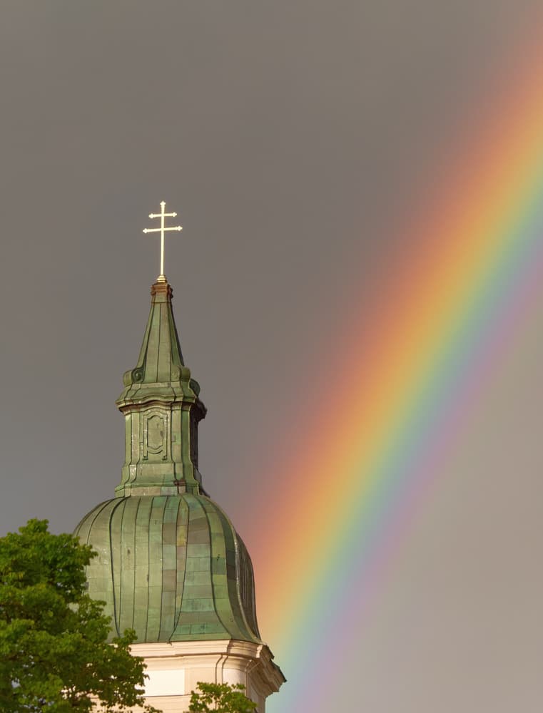 A church steeple with a cross atop under a cloudy sky with a bright rainbow beside it.