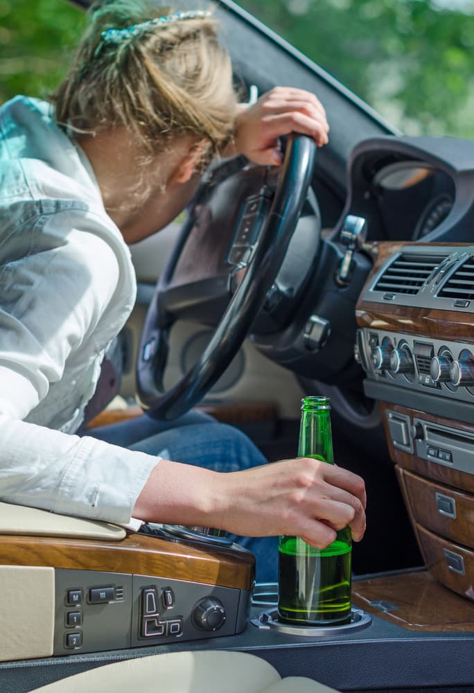 A woman rests their head on a steering wheel, holding a beer bottle, implying impaired driving.
