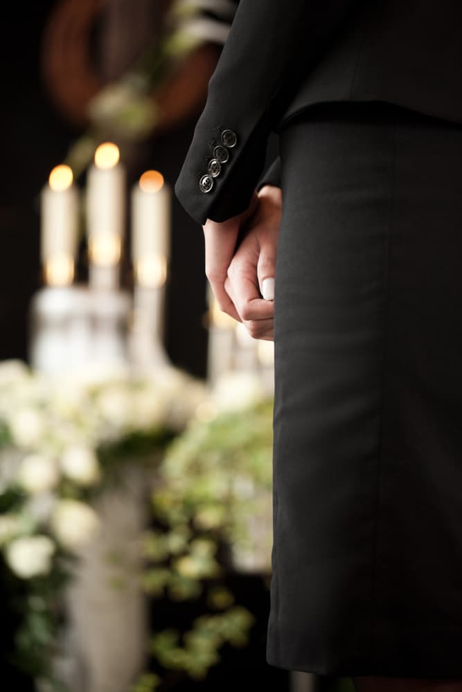 A solemn individual at a funeral, hands clasped, with candles and flowers in soft focus.