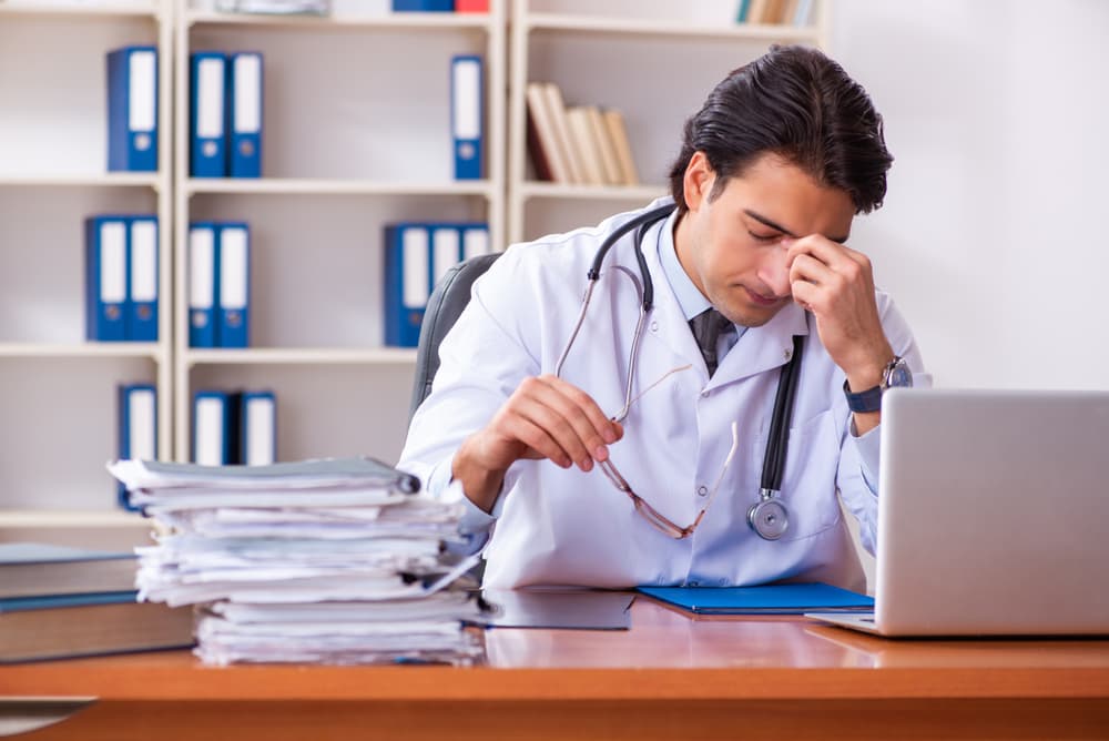A doctor with a stethoscope looking tired and stressed in front of a computer and papers.