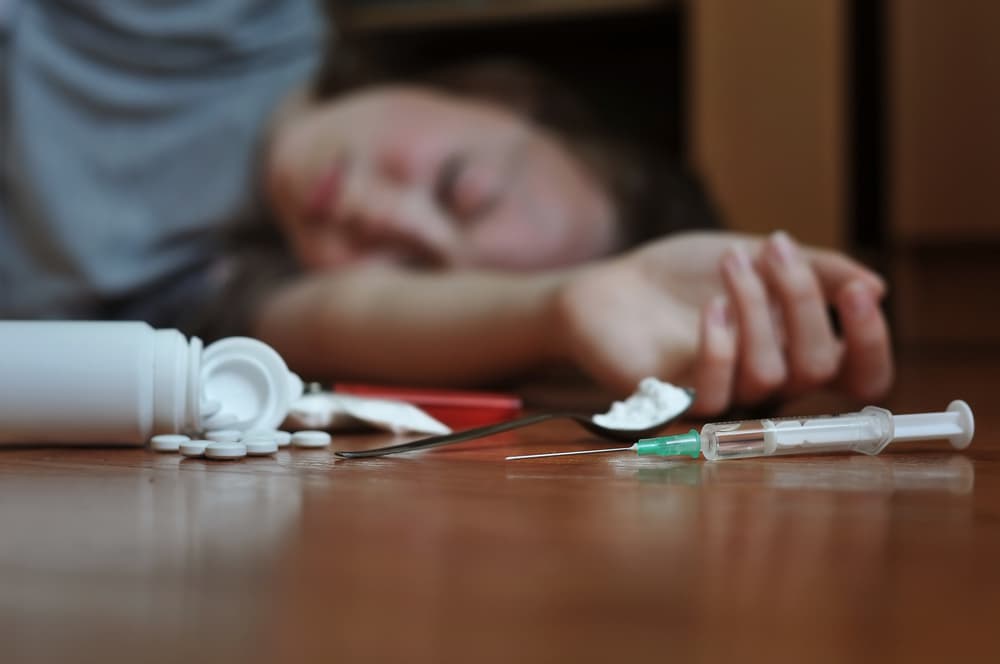 A person who is unconscious with a spill of pills from a bottle and a syringe nearby.