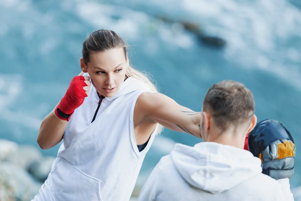 Woman in white practicing boxing punches with a partner wearing focus mitts by the sea.