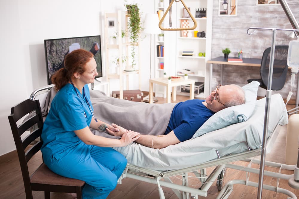 Elderly man with health issues lying in a nursing home bed, supported by a compassionate female doctor providing care.