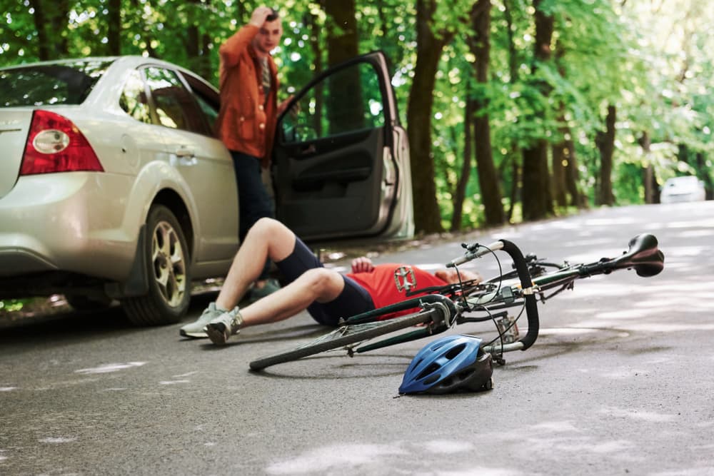 A bicycle and a silver car collide on a forest road during the daytime.