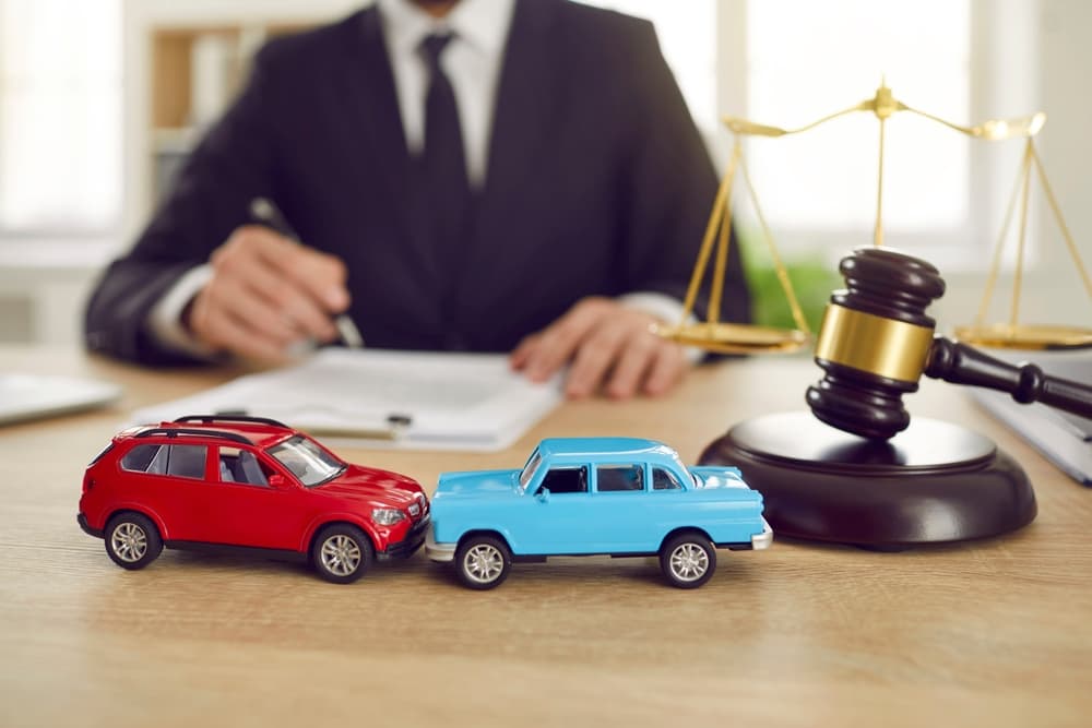 A close-up view shows two toy model cars depicting a collision, set on a table in front of a judge overseeing a car accident lawsuit. Beside the cars are a judge's gavel and the scales of justice, symbolizing the legal proceedings.