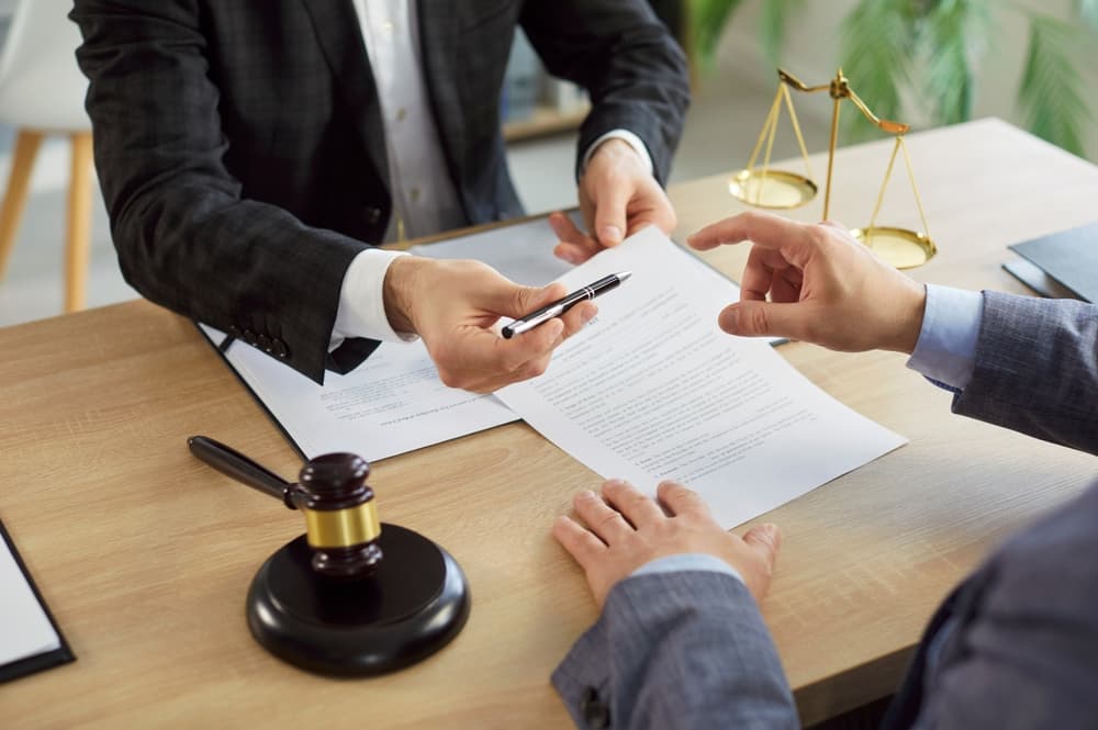 A man signs a contract while consulting with a male advocate in an office setting. The lawyer, seated at a desk with a judge's gavel and scales of justice, provides legal guidance during the meeting.