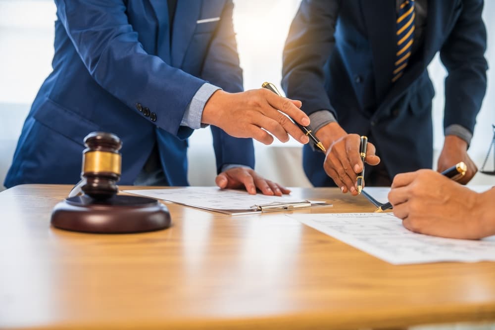 A group of business professionals and a lawyer in formal attire meet around a conference table, reviewing a contract and discussing legal and litigation matters.