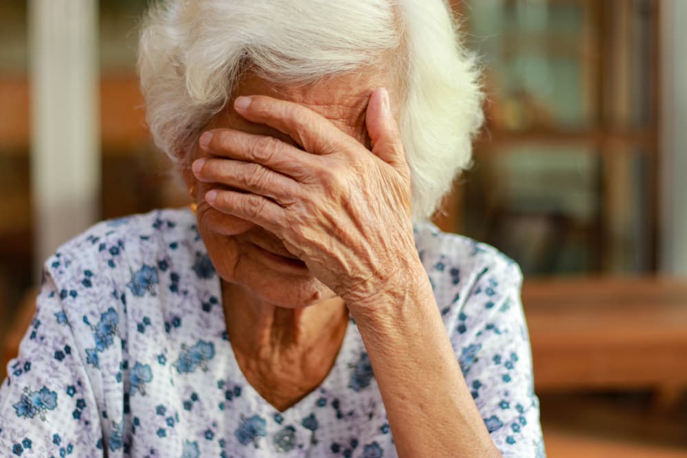 An elderly Asian woman with white hair shows signs of memory impairment. She faces ongoing health problems and rising treatment costs.