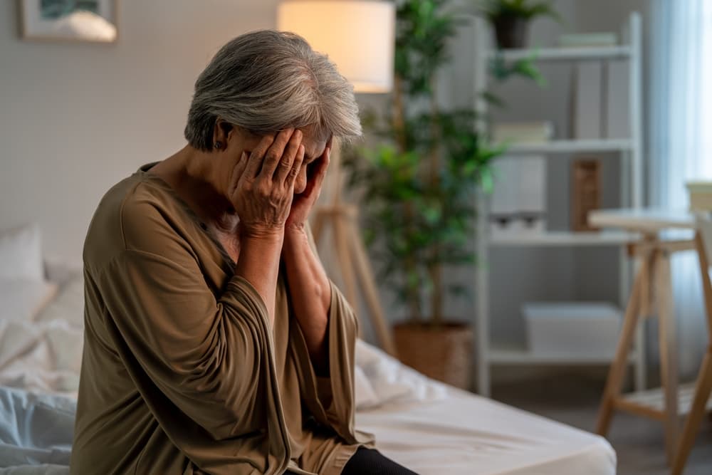 A senior woman sits alone in her bedroom, feeling sad and frustrated while thinking about financial stress, nursing home abuse, and life challenges.