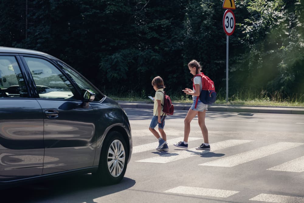 A car approaches children crossing the street after school, while the kids are distracted, looking at their smartphones.