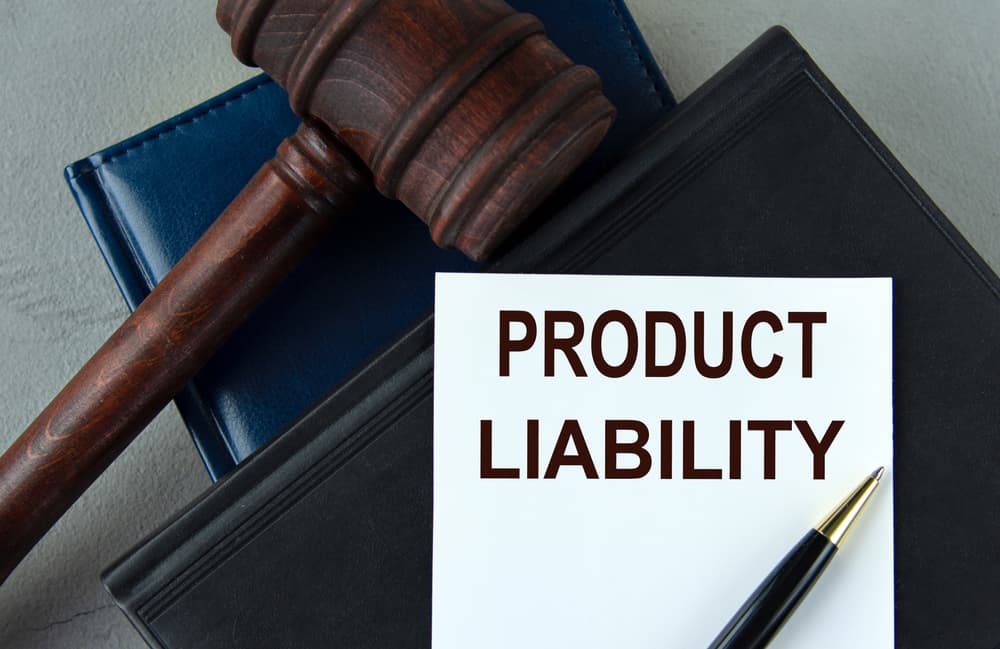 The words "Product Liability" appear on a white sheet alongside leather notebooks, a judge’s gavel, and a pen, symbolizing legal documentation and justice.