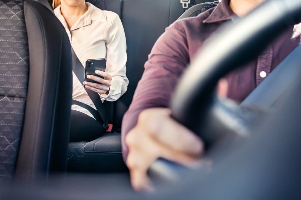 Woman using smartphone in the back seat of a car or taxi. Rideshare passenger with driver visible. Mobile app in use during ride.