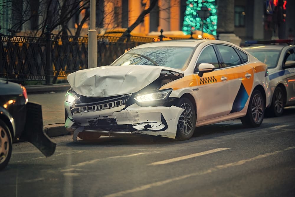 Evening car crash scene involving a taxi. The vehicle sits roadside with a severely damaged front end, showing clear signs of a recent collision.