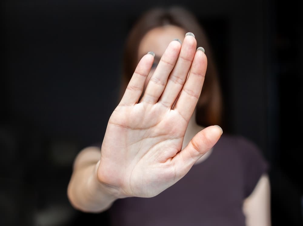 Close-up of a determined young woman holding up her hand in a stop gesture toward the camera. Blurred background. She stands against abuse, bullying, and gender discrimination.