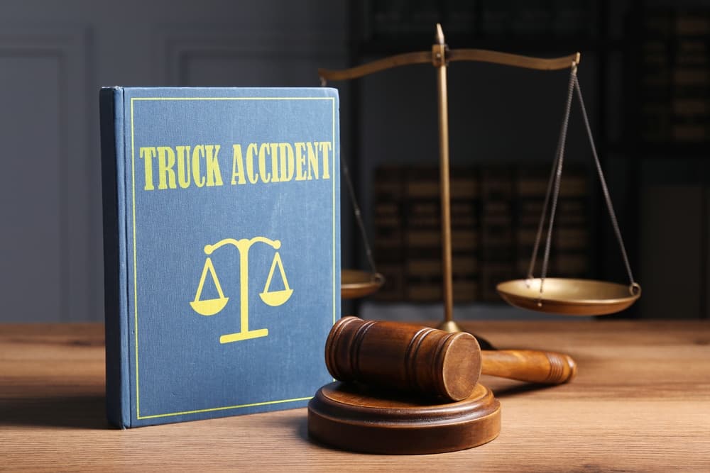 Truck accident law book, judge’s gavel, and justice scales arranged on a wooden table in a courtroom setting.
