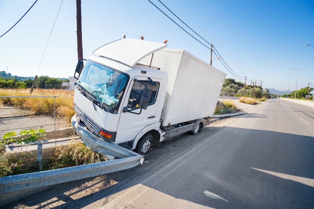 Truck crash scene with shattered windshield and damaged road barrier under clear, sunny skies.