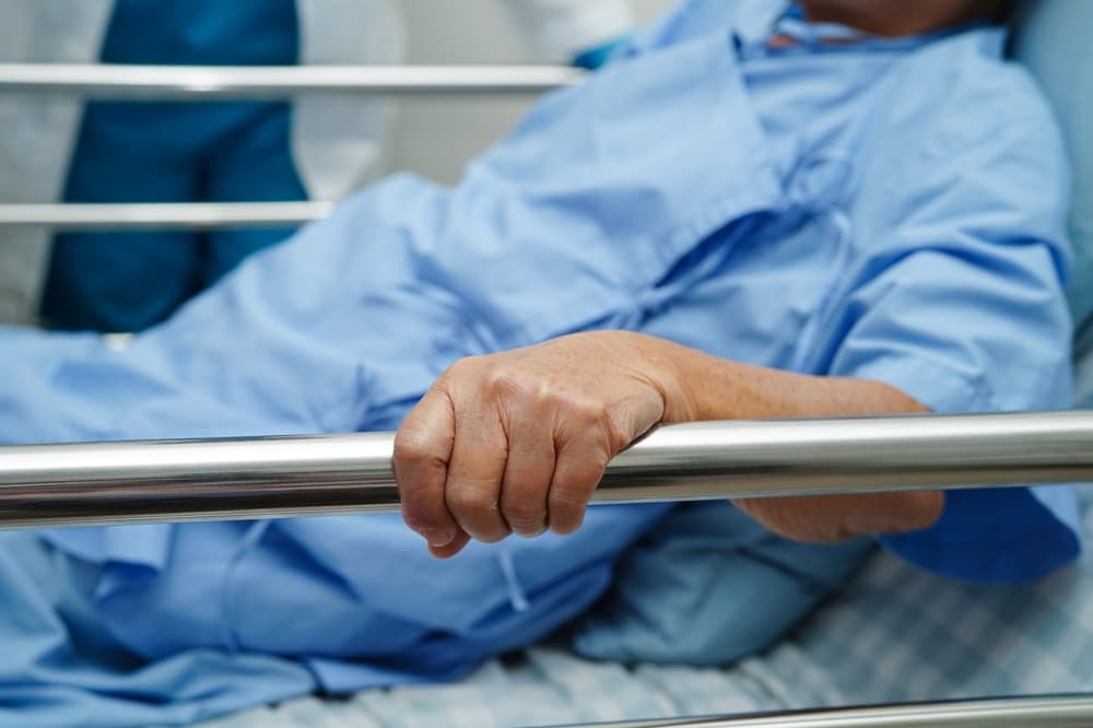 Asian elderly woman lies in a hospital bed, gently holding the bed rail, gazing with hope as she waits for her family to arrive.