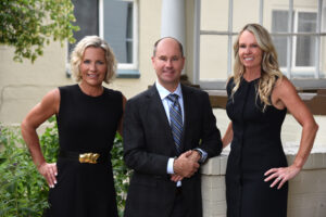 Top Dog Law Sexual Assault Team — two women in black dresses and one man in a dark suit with a striped tie, standing together outside a building, smiling at the camera.
