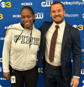 Top Dog Law attorney in a navy suit with a red tie standing beside a smiling woman in a grey 'PINK' hoodie, posing together in front of a CBS Philly and CW branded backdrop.